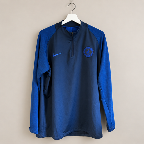 A blue and royal blue long-sleeve Nike training top with a quarter-zip collar, hanging on a white hanger against a plain beige wall. The Chelsea FC crest is embroidered on the left chest, and the Nike swoosh is on the right.
