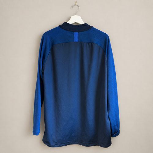 A long-sleeve athletic jersey shown from the back, featuring a dark navy blue main body and contrasting royal blue sleeves and collar, hanging on a white hanger against a plain beige wall.