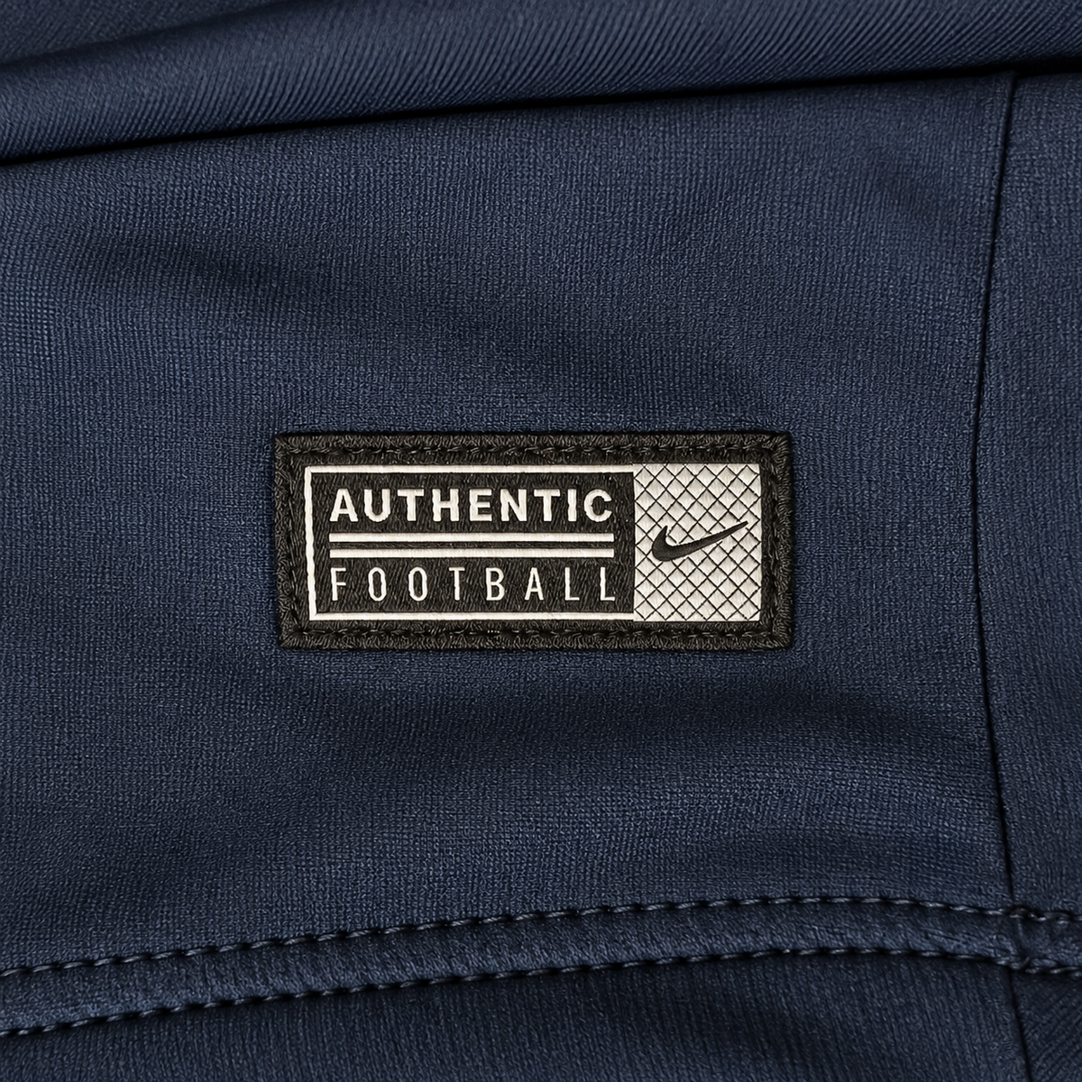A close-up of a navy blue athletic jacket showing a rectangular embroidered patch on the chest. The patch is black and off-white with the text 'AUTHENTIC FOOTBALL' and a small Nike swoosh.
