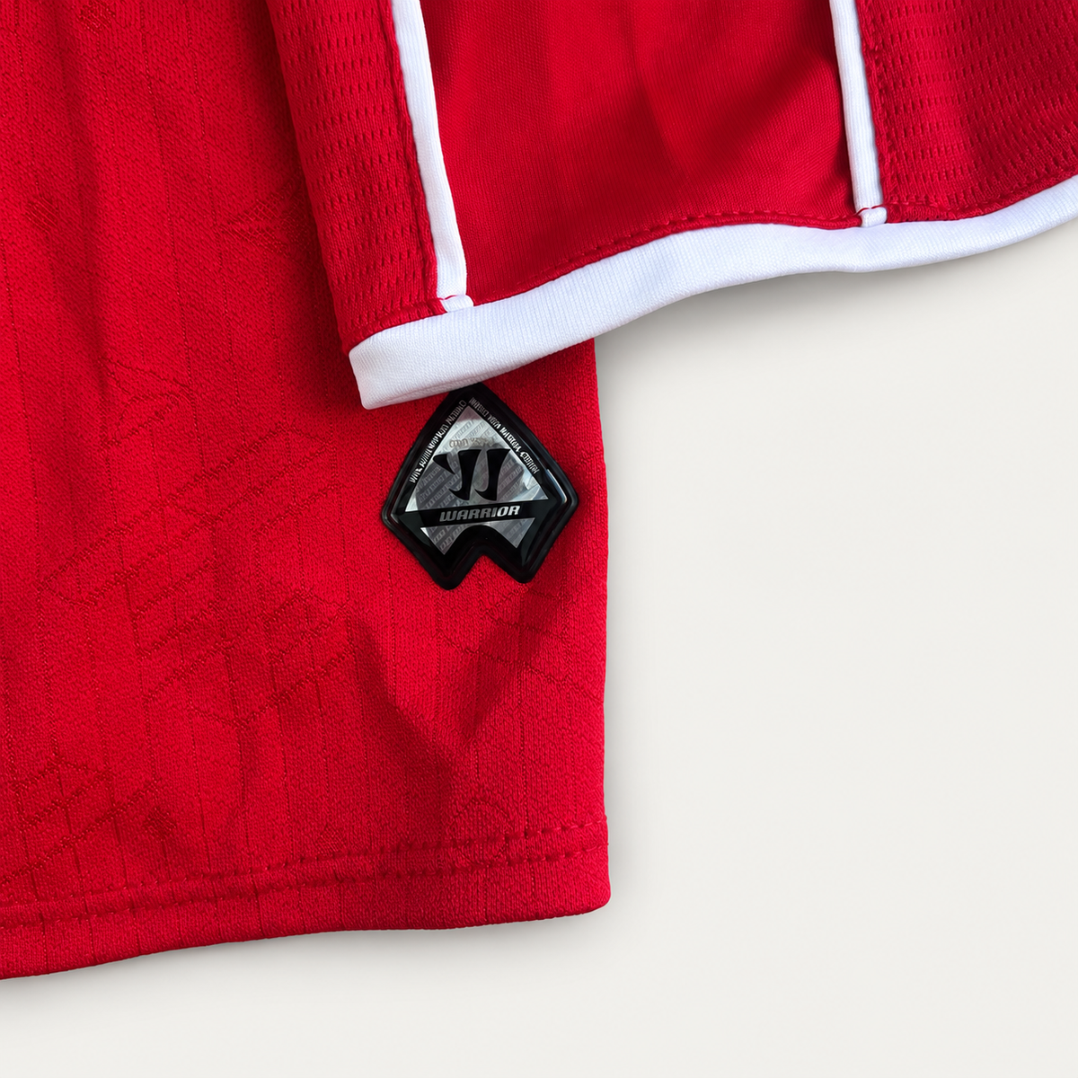 Close-up of a red athletic jersey or shorts with white piping and a black-and-gray triangular brand patch near the hem, shown on a light neutral background.