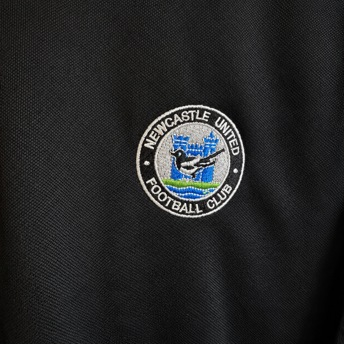 Close-up of a black garment interior showing a black hangtag and a black brand label with gold foil branding. The hangtag reads 'SD74' and 'UNIC DIRECT EXCLUSIVE', and the label reads 'Aussie Retro Official Affinity'.