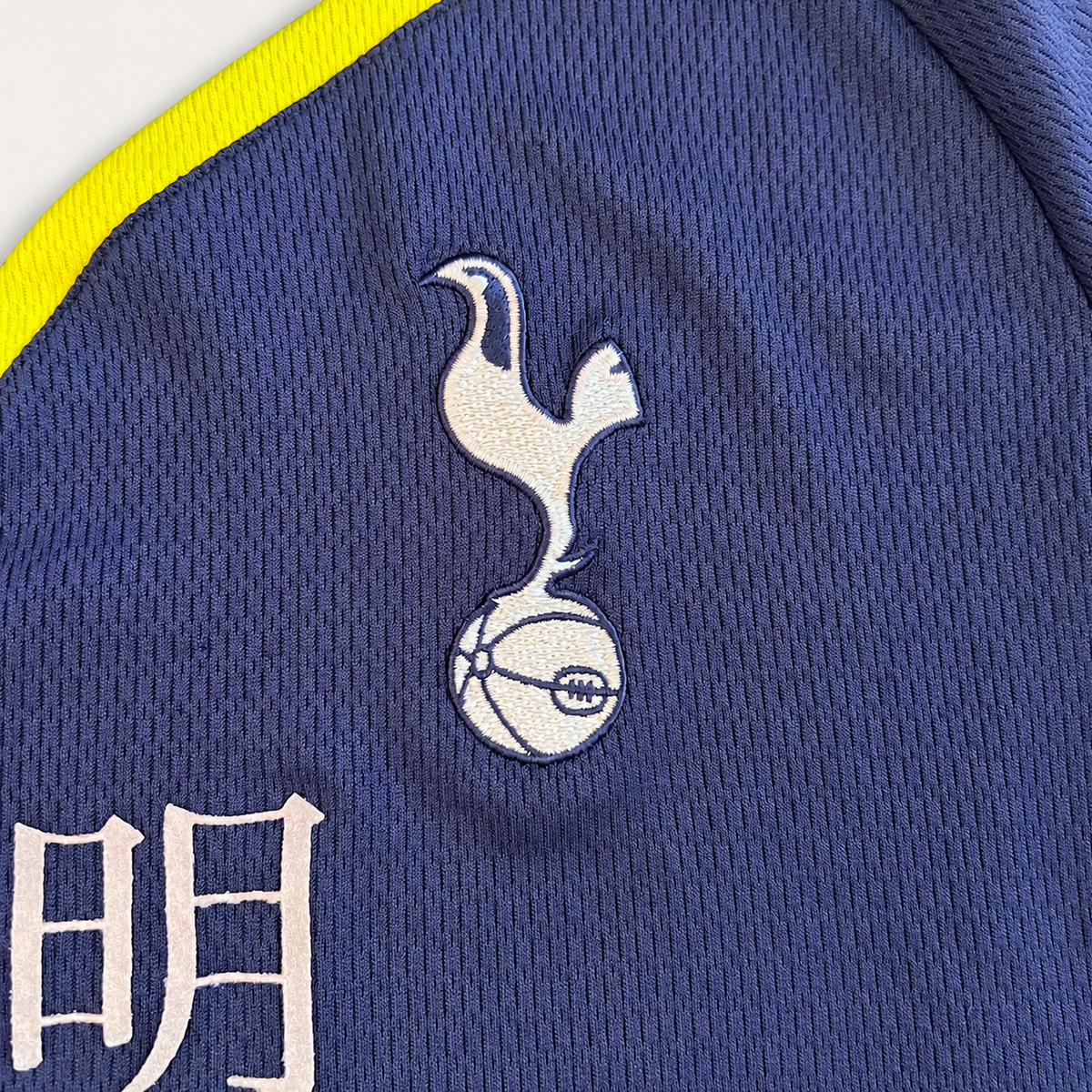 Close-up of a navy blue athletic jersey fabric with a white embroidered Tottenham Hotspur cockerel logo and football motif, plus a bright yellow trim along the edge.