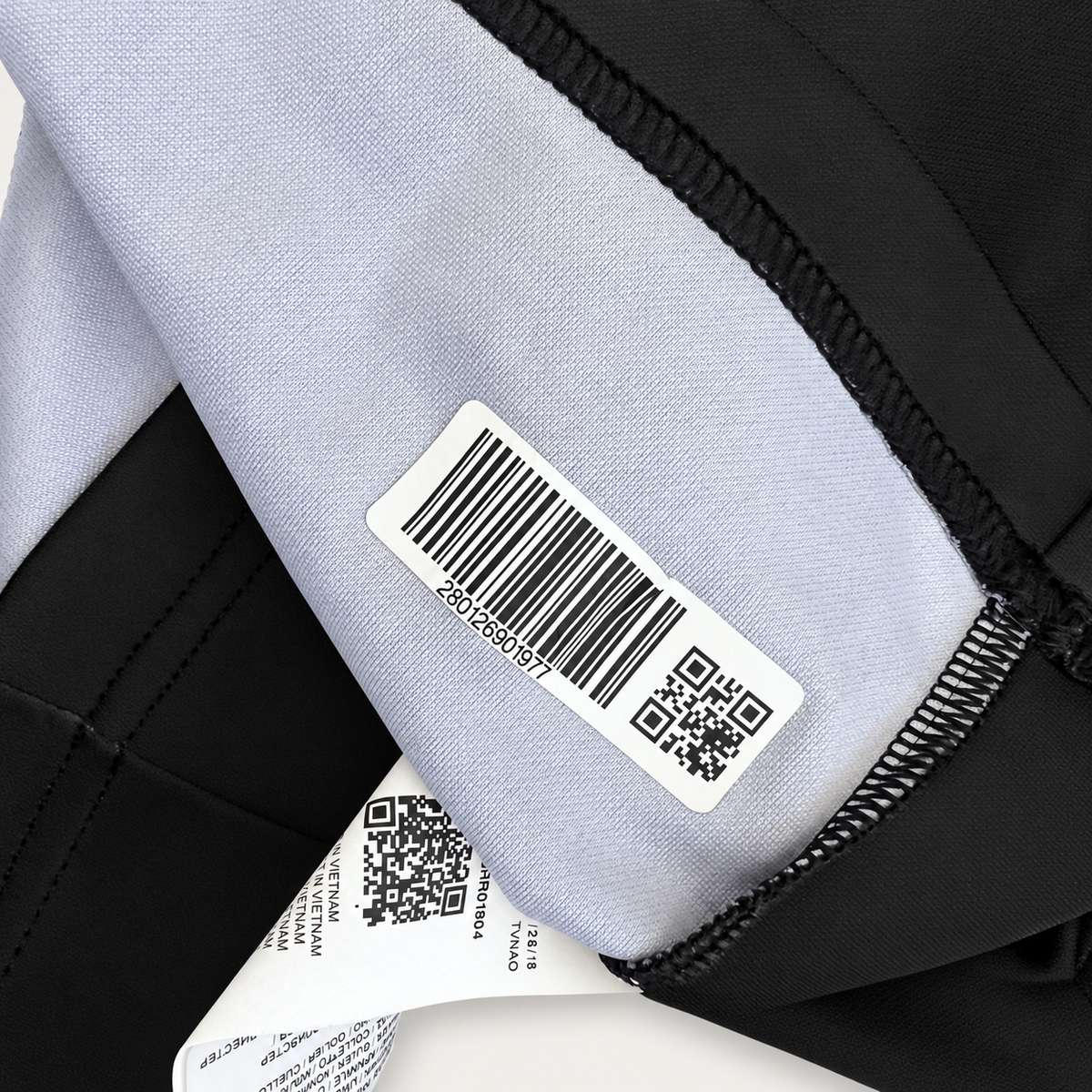 Close-up of a garment's interior label area showing a white barcode sticker and a sewn-in product tag on black and light gray fabric.