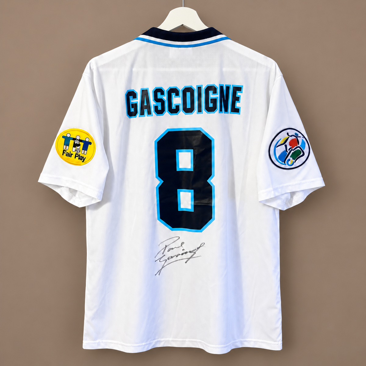A white vintage football jersey with 'GASCOIGNE' and the number '8' printed on the back in navy and light blue, hanging on a white hanger against a plain brown wall. It features a yellow 'Fair Play' patch on the right sleeve and a circular patch on the left sleeve.