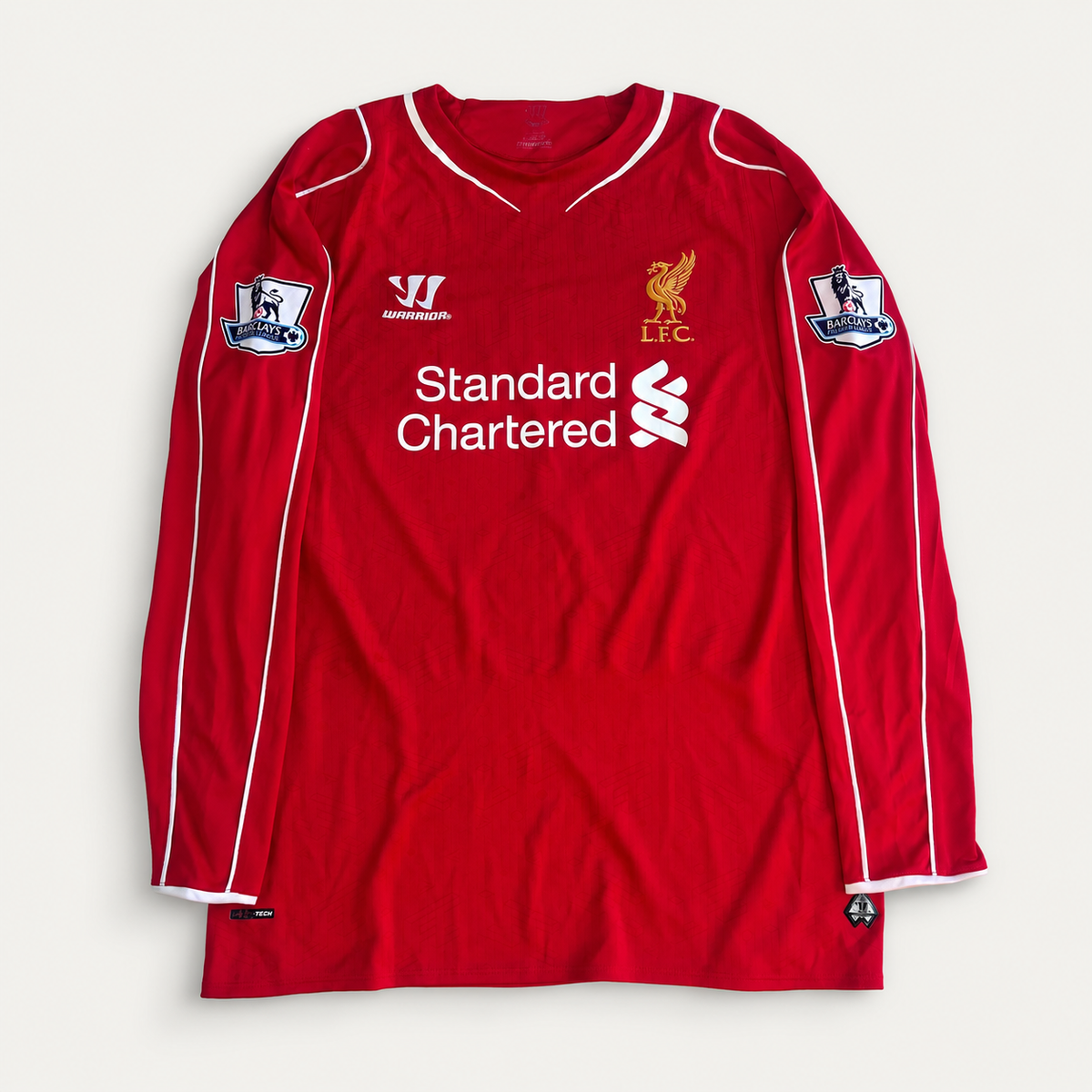 A red long-sleeve Liverpool football jersey with white trim, featuring the 'Standard Chartered' sponsor logo on the chest, the Wacker logo, and the LFC crest. It includes sleeve patches and is laid flat against a light off-white background.